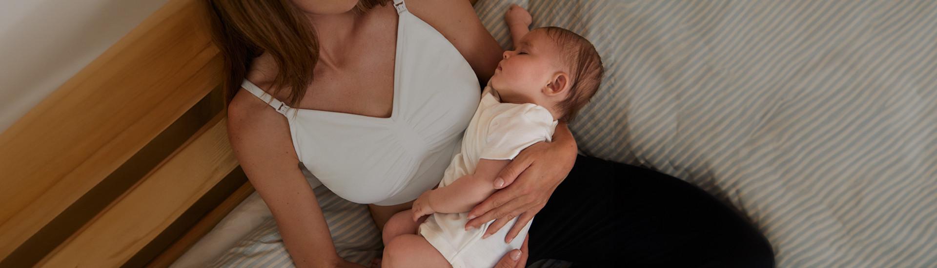 Woman in white nursing bra holds sleeping baby wearing white bodysuit on striped bedding.