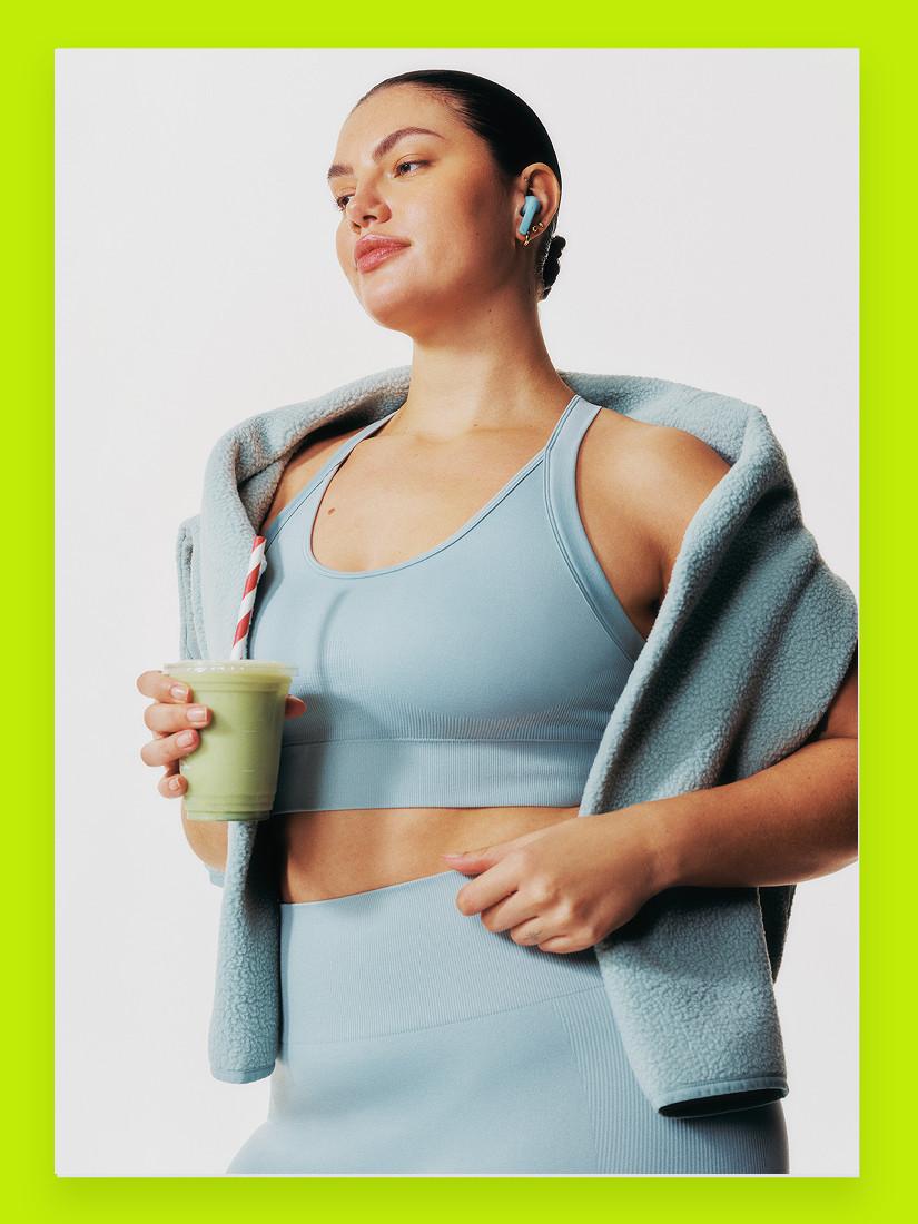 Person wearing blue sports bra and matching leggings with fleece jacket, holding green drink and wireless earbuds.