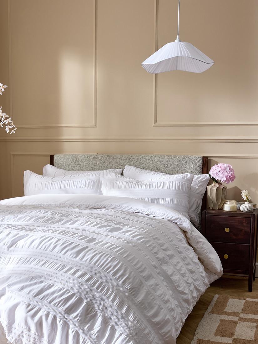 Elegant bedroom featuring white textured bedding, pleated pendant light, and wooden bedside table with pink flowers.