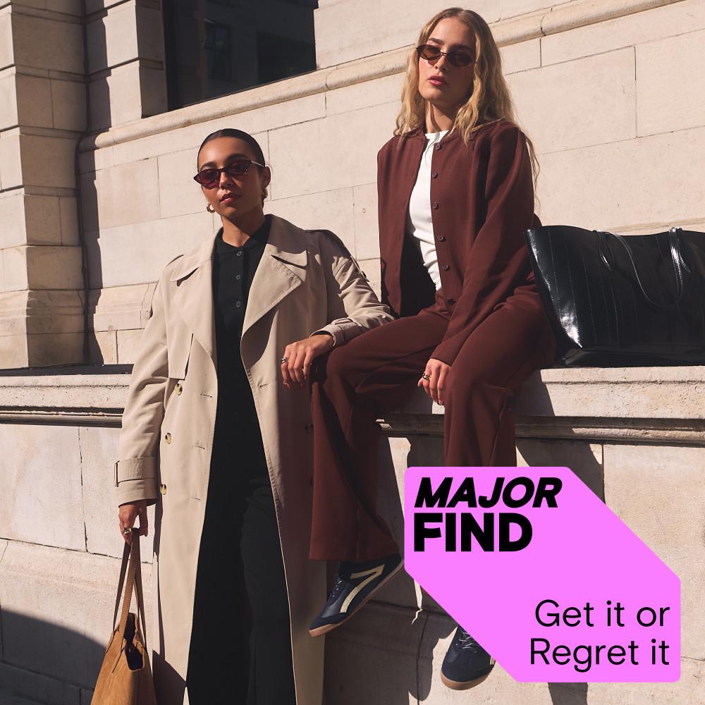 Two models wearing sunglasses and tailored outerwear sitting on stone steps in urban setting.