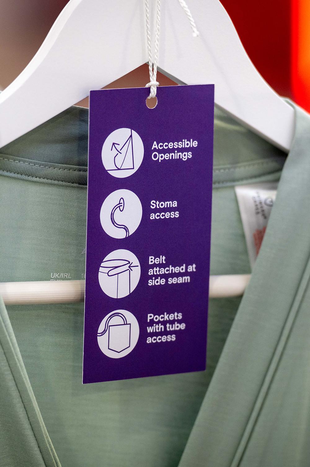 Purple accessibility tag on sage green garment showing icons for accessible openings, stoma access, belt attached at side seam and pockets with tube access.