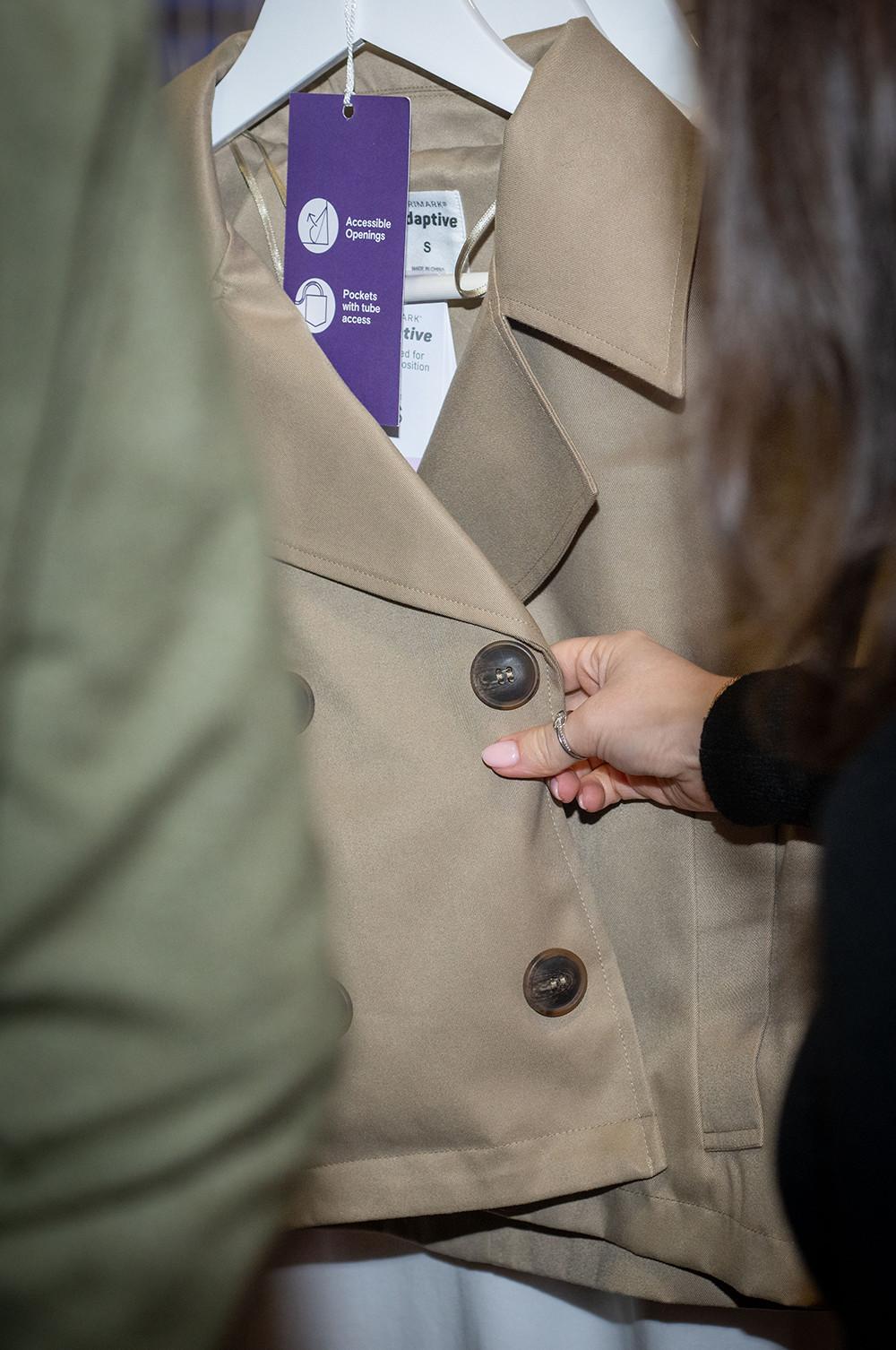Hand fastening button on beige trench coat with purple accessibility tag showing adaptive clothing features.