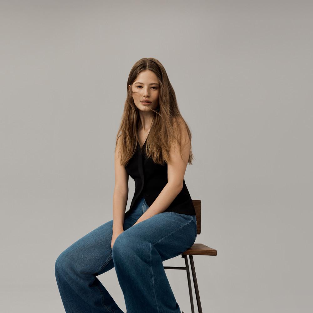 Person wearing black sleeveless top and blue jeans sitting on wooden chair against neutral backdrop.
