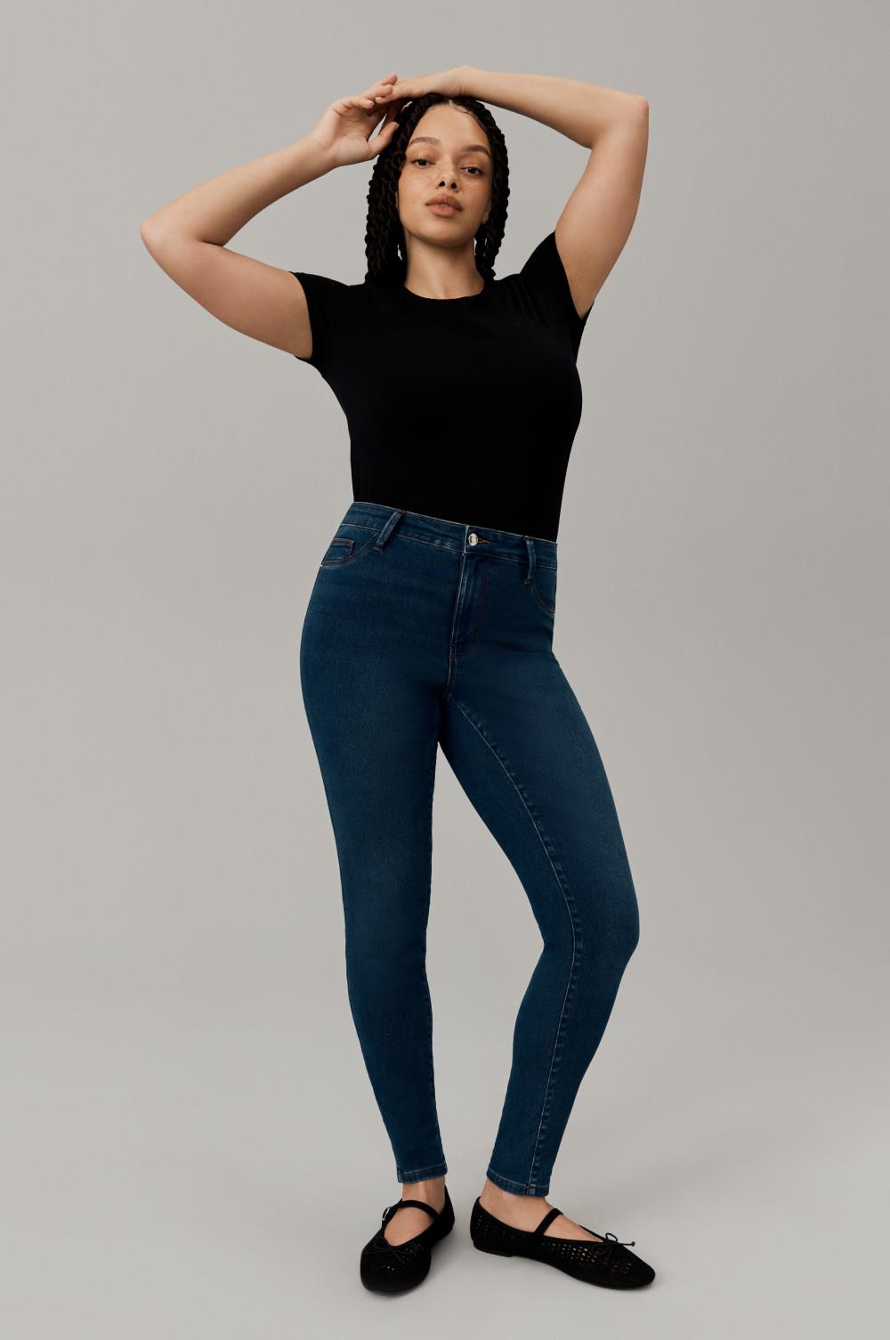 Person wearing black fitted t-shirt with dark blue skinny jeans and black ballet flats, posing with arms raised.
