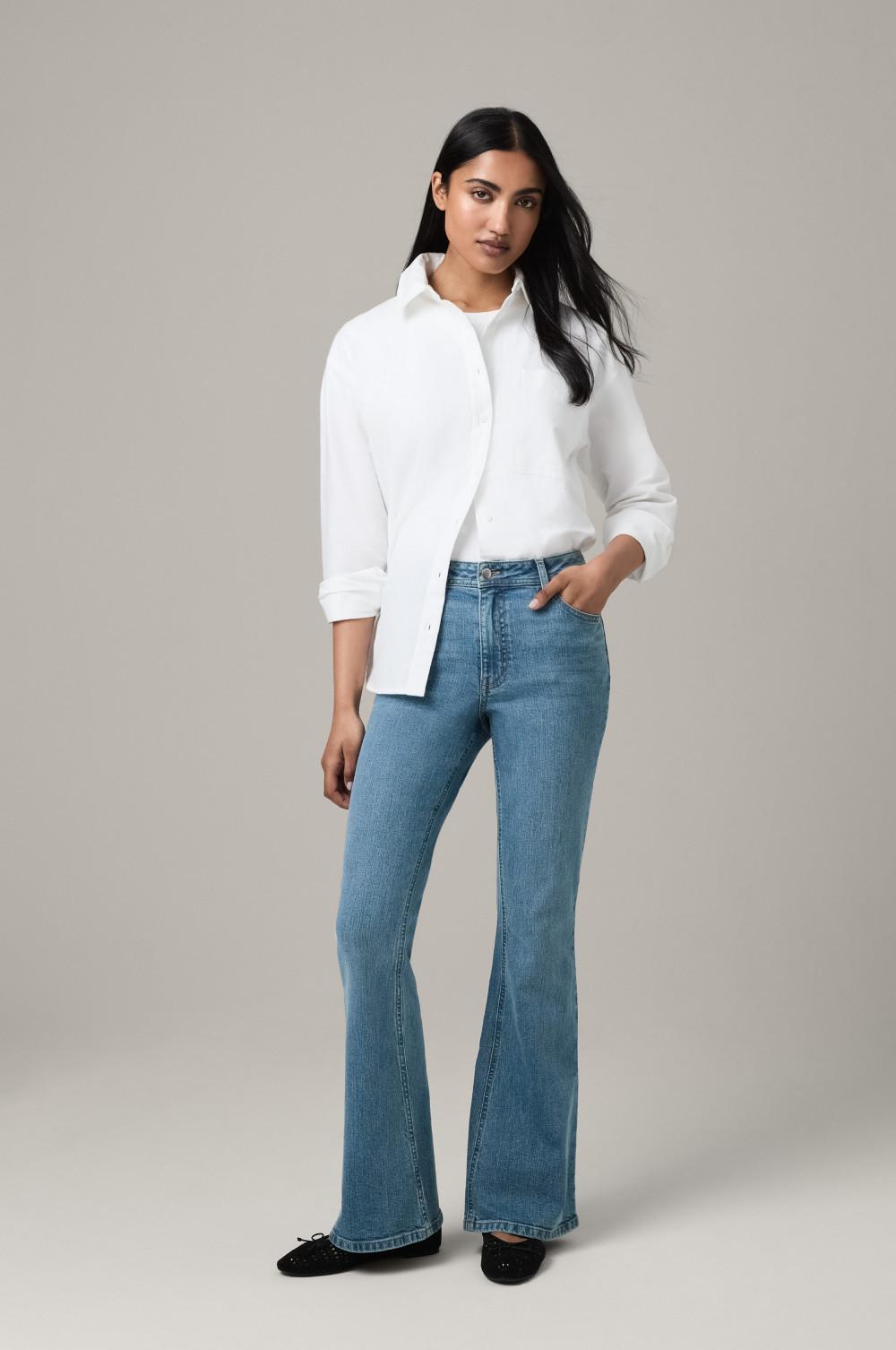 Person wearing classic white button-up shirt with blue flared jeans and black shoes against neutral backdrop.