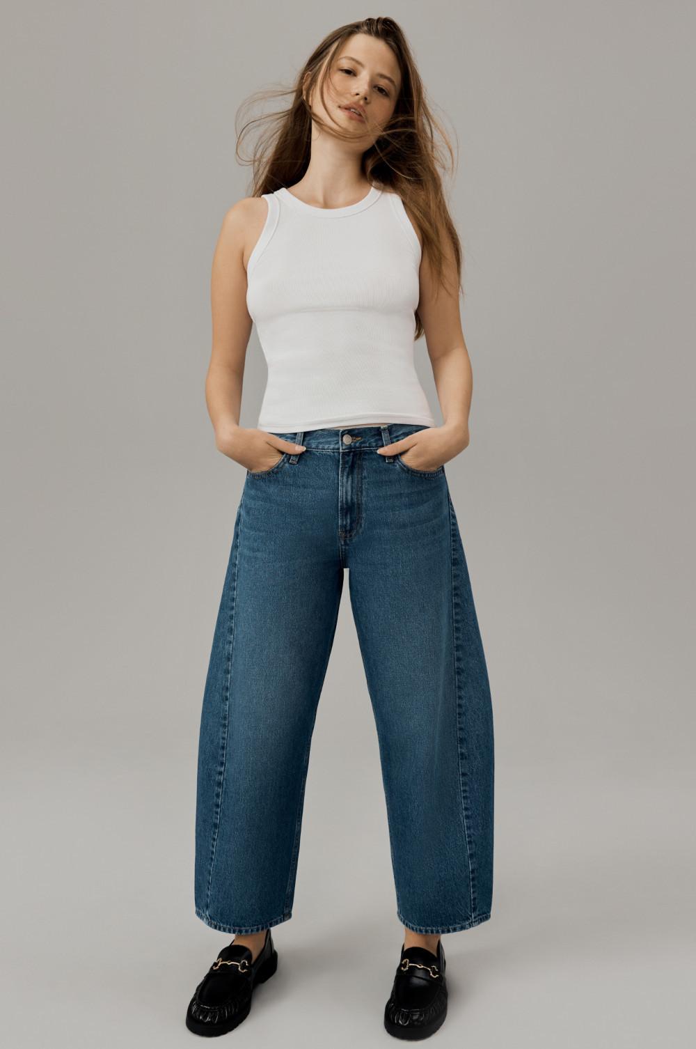 Person wearing white ribbed tank top with wide-leg blue jeans and black loafers against neutral background.