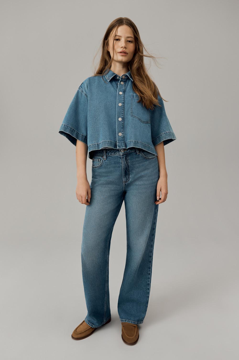 Person wearing cropped denim shirt with short sleeves paired with wide-leg jeans and brown loafers.