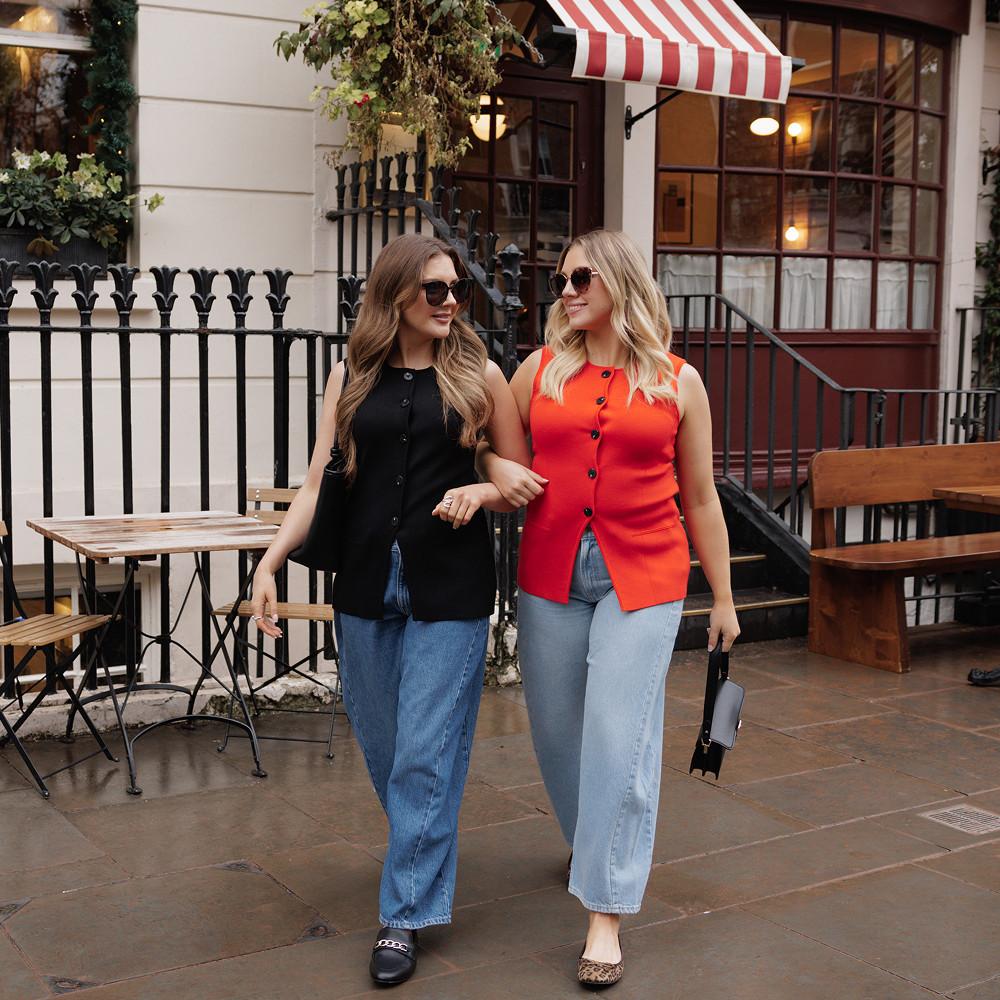 Dve ženski, ki nosita sončna očala, hodita z roko pod roko pred kavarno, oblečeni v majice brez rokavov z gumbi in kavbojke.