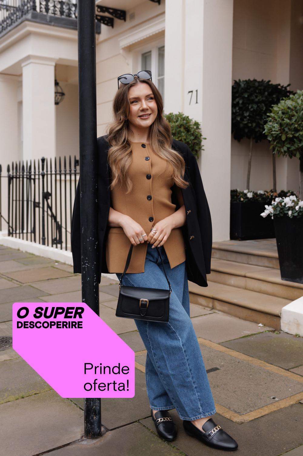 Persoană care poartă un cardigan maro camel cu nasturi, un palton negru, blugi albaștri și mocasini negri, cu o geantă mică neagră, în fața unei case.