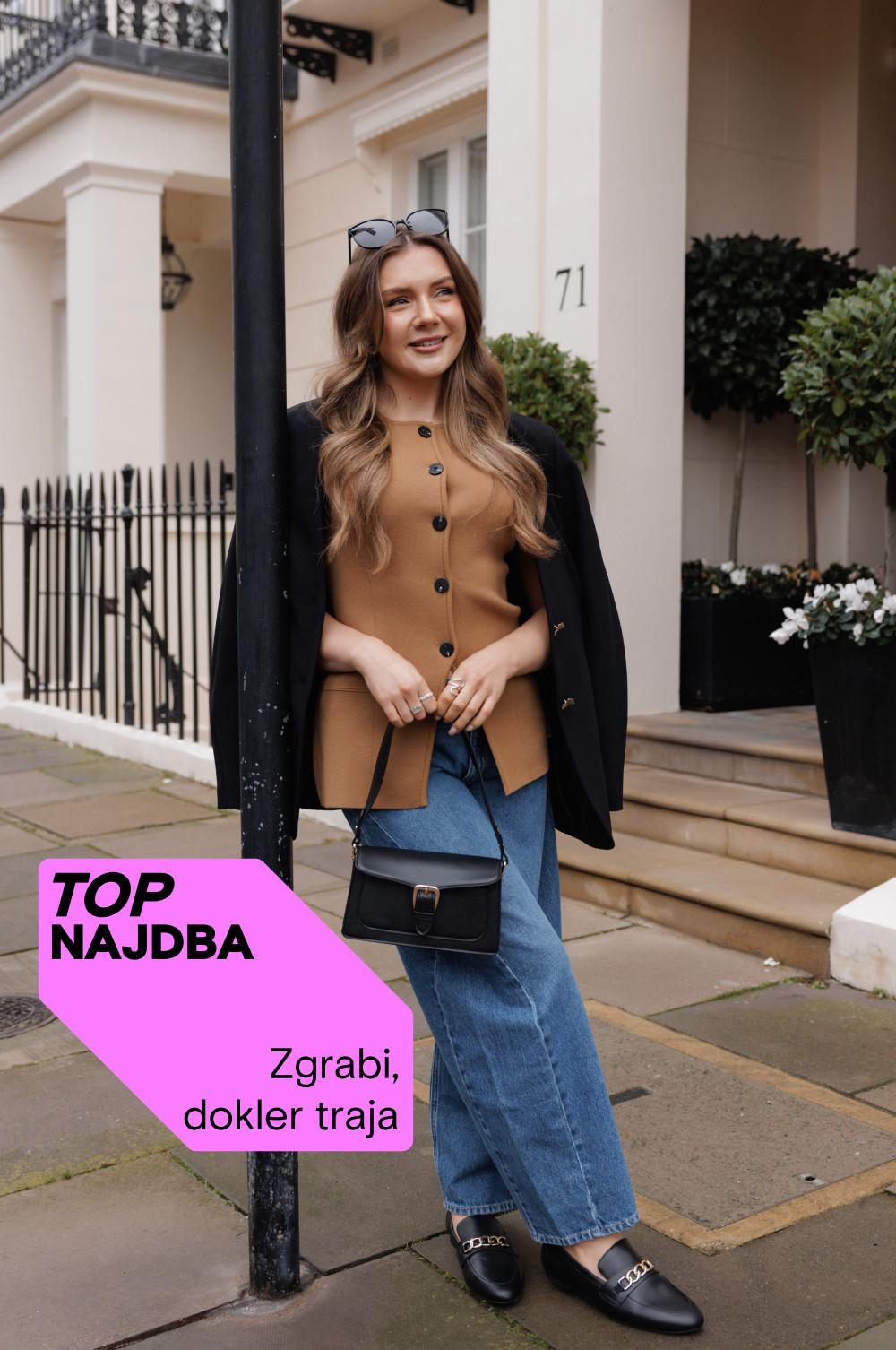 Oseba, oblečena v kameljo jopico z gumbi, črn plašč, modre kavbojke in črne mokasine z majhno črno torbico pred vrstno hišo.