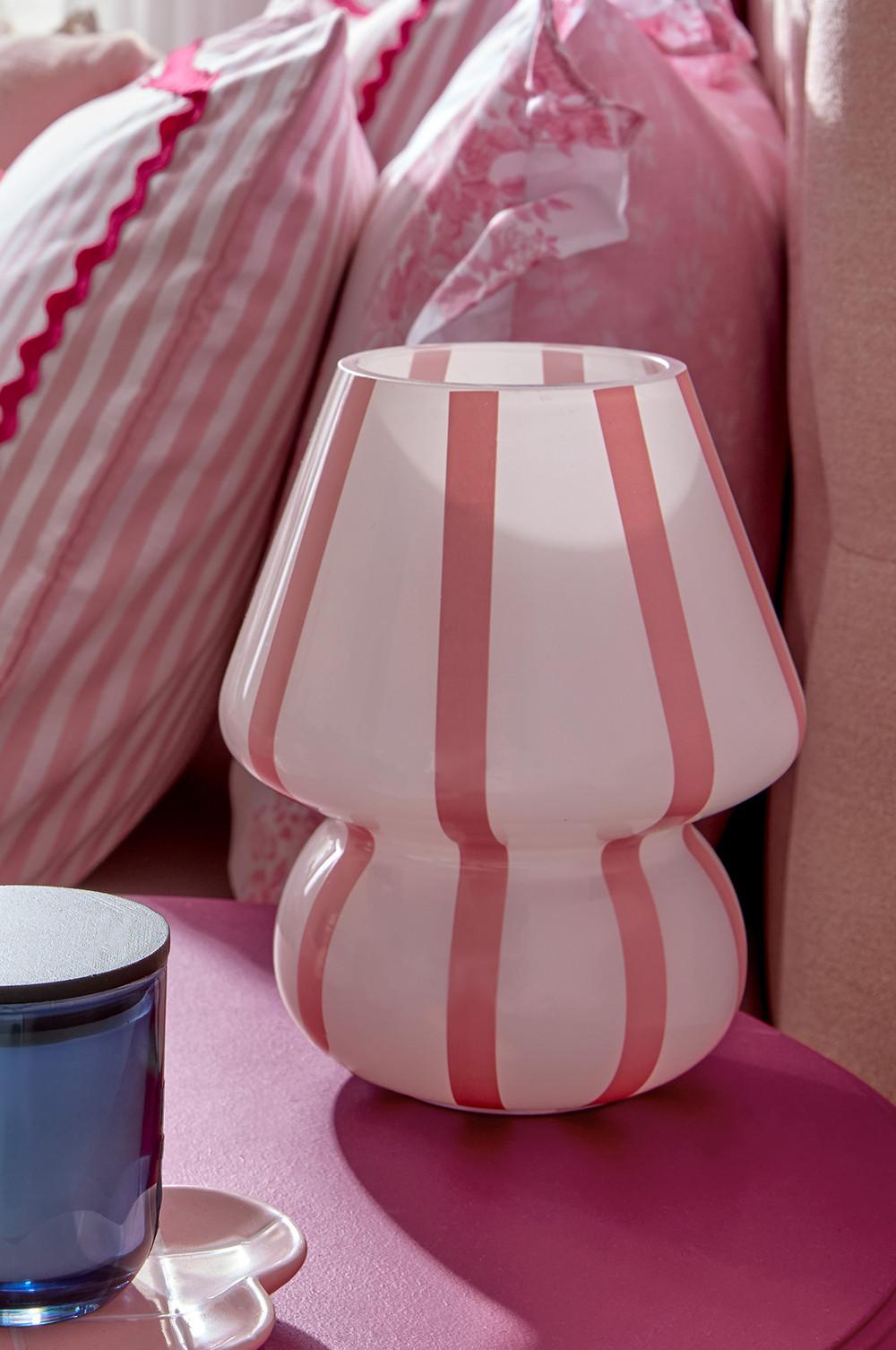 Glass lamp with pink and white vertical stripes, curved hourglass shape, on pink surface with pillows behind.