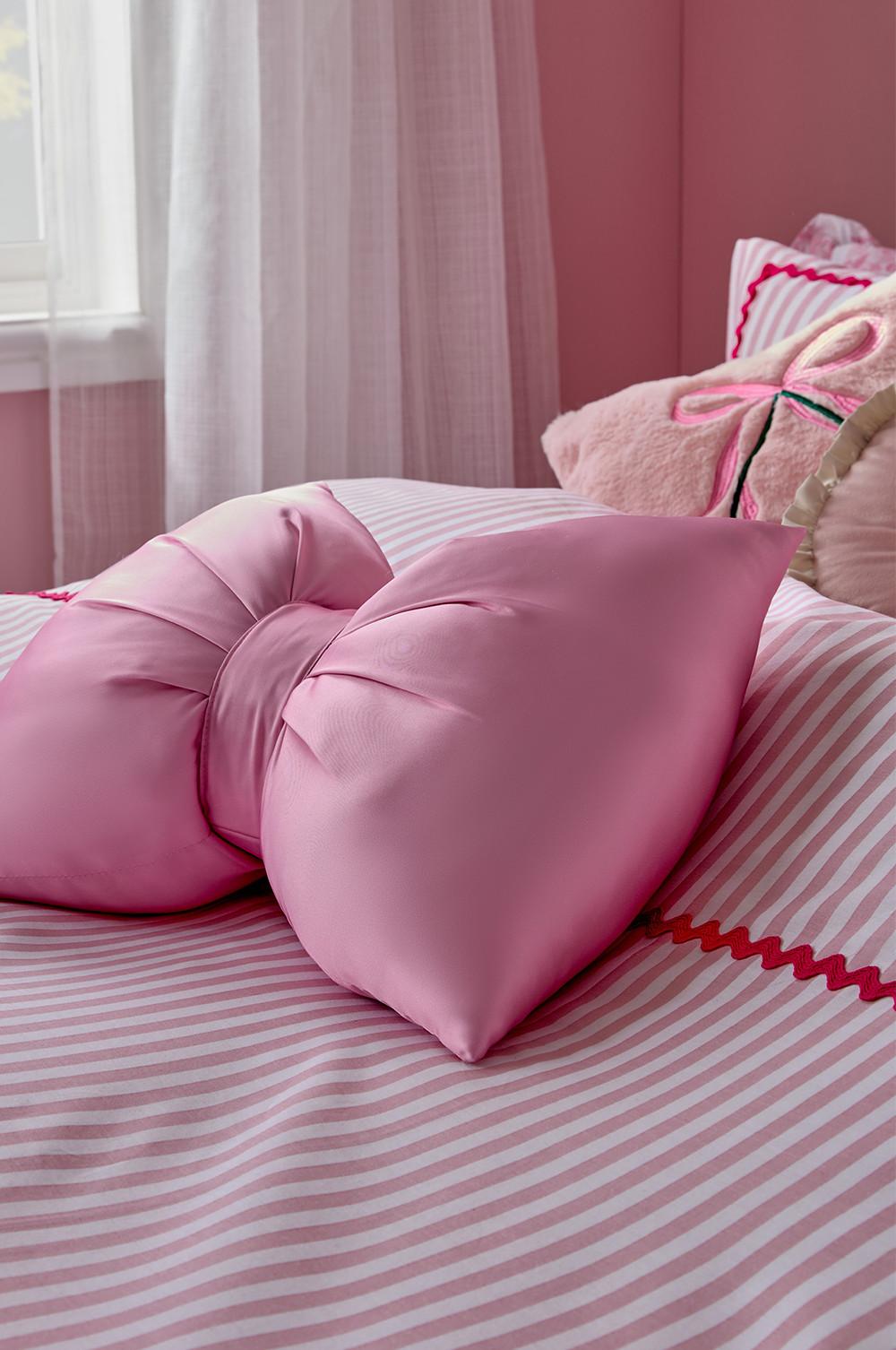 Pink bow-shaped decorative cushions on striped bedding with white curtains and cherry-detailed pillows in background.