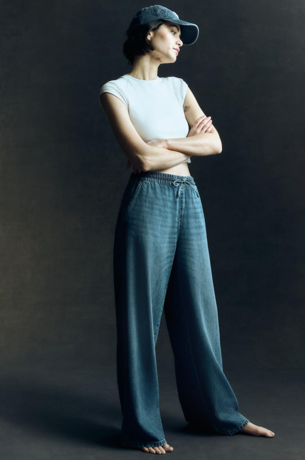 Wide leg denim joggers, white cropped T-shirt, blue baseball cap worn with arms crossed.