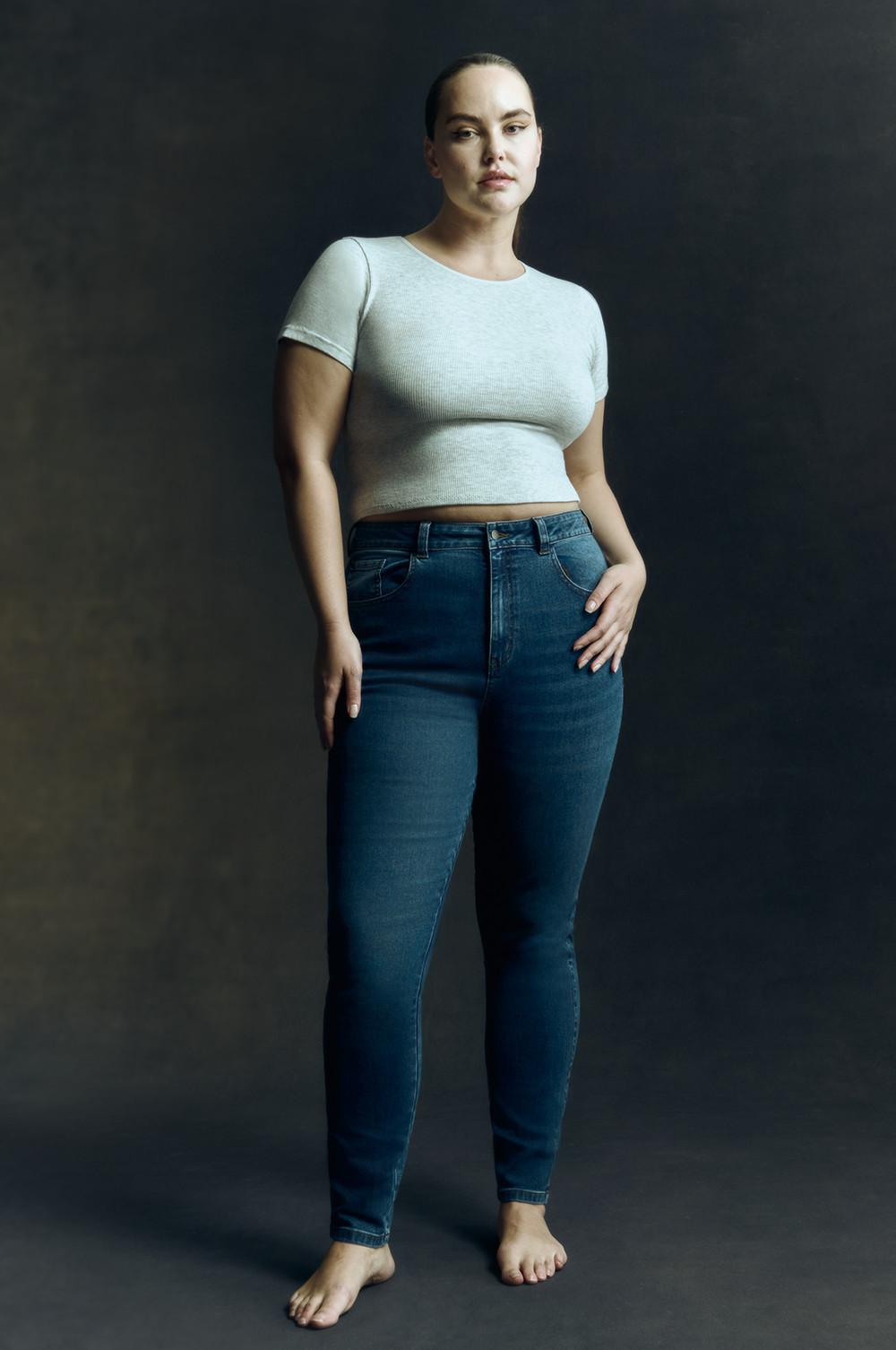 Model in white cropped tee, high waist blue skinny jeans, barefoot on dark backdrop.