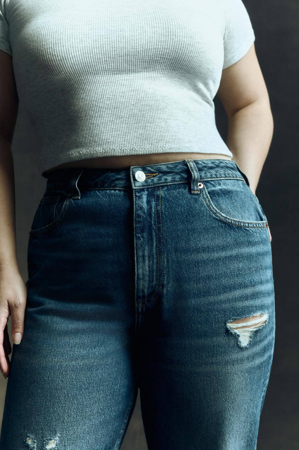 Nahaufnahme einer blauen Mom-Jeans im Used-Look mit zerrissenen Knien, getragen zu einem kurzen weißen Rippen-T-Shirt.