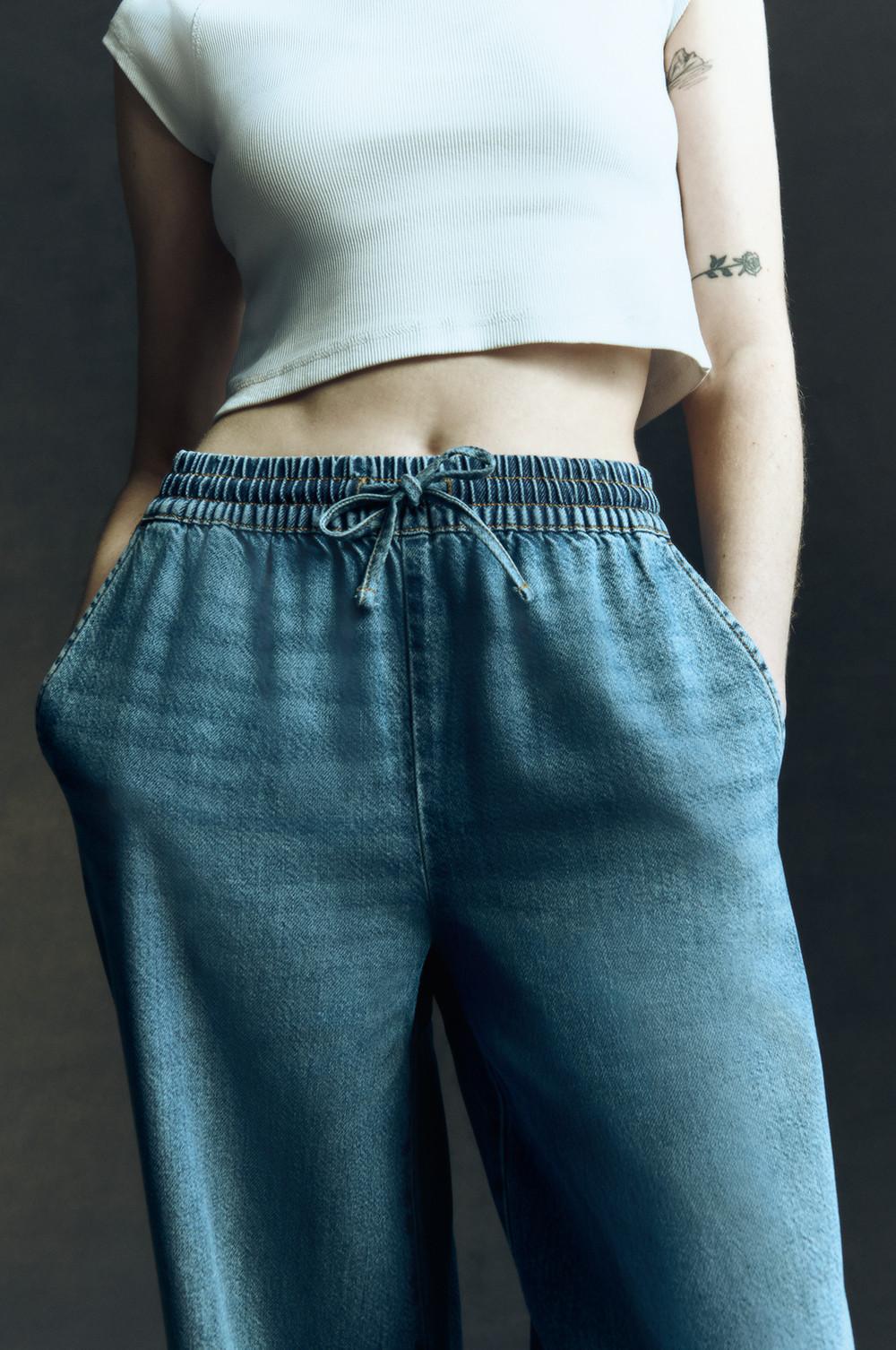 Model in blauer High-Waist-Jeans mit weitem Bein und weißem bauchfreiem Top.