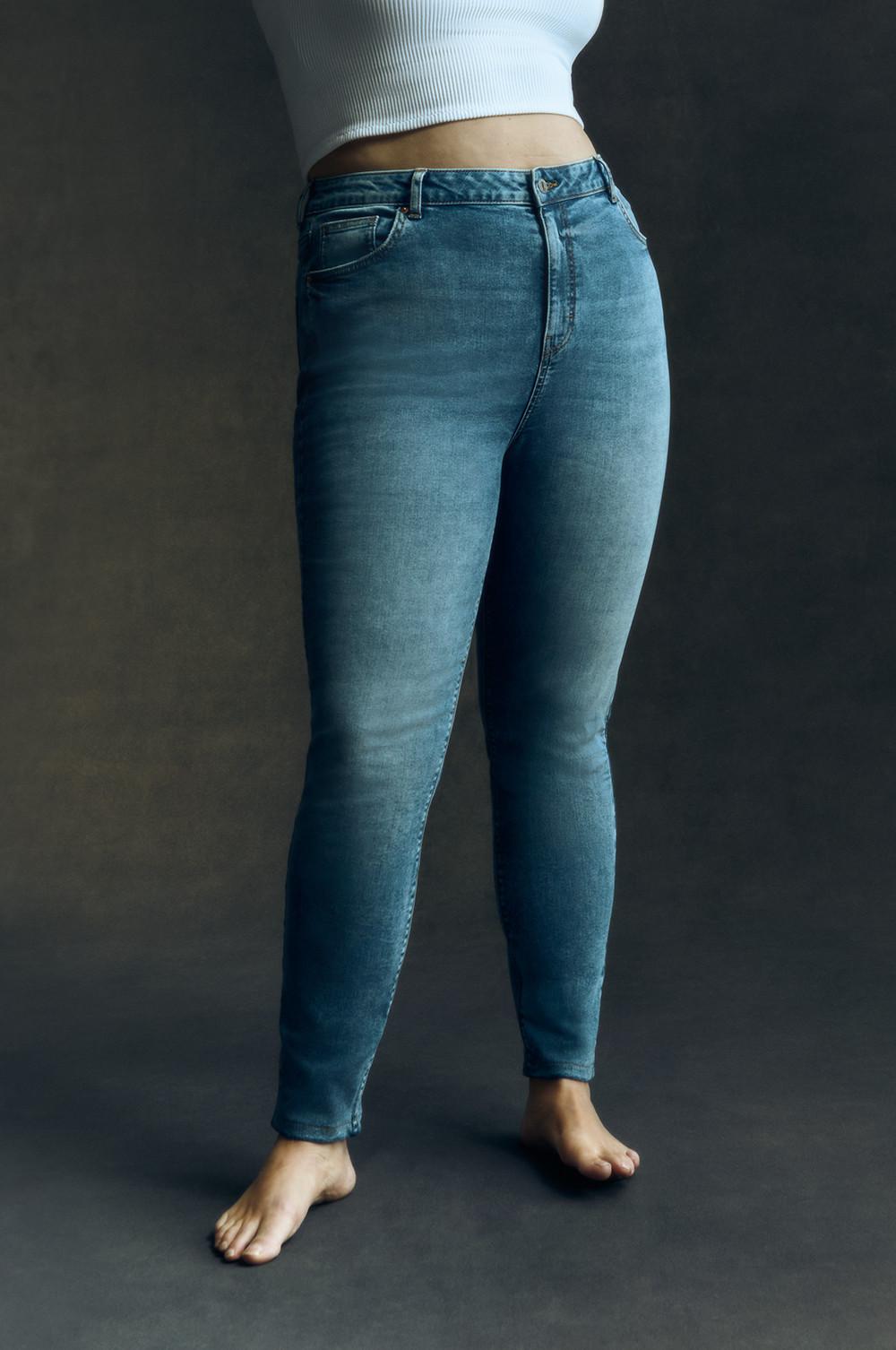 Das Model trägt eine blaue Skinny-Jeans mit hoher Taille und ein weißes Top und posiert barfuß stehend.