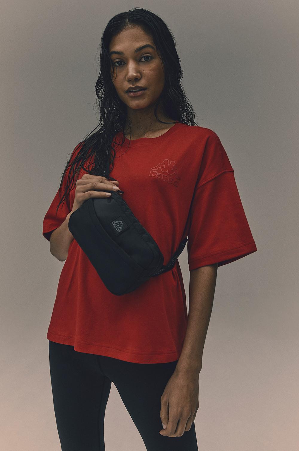 Das Model trägt ein rotes Kappa-T-Shirt mit Logo, eine schwarze Umhängetasche und eine schwarze Hose.