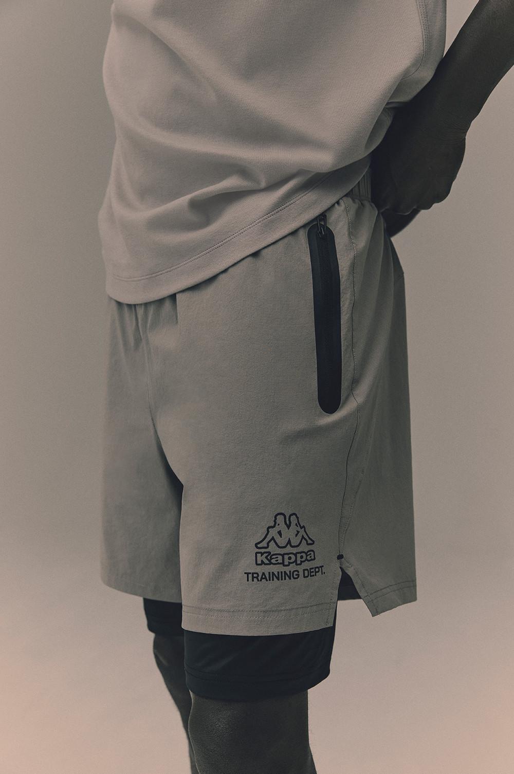 Nahaufnahme einer grauen Kappa Training Dept. Sporthose mit schwarzer Reißverschlusstasche an der Seite und Logo-Print am Bein sowie einem weißen T-Shirt