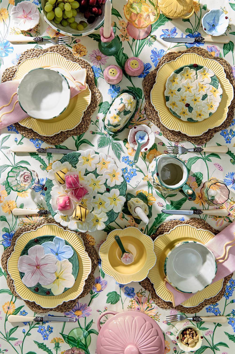Spring table setting with floral plates on woven placemats, and pink candles, yellow bowls, pink ramekin and green floral cake stand on a floral tablecloth.