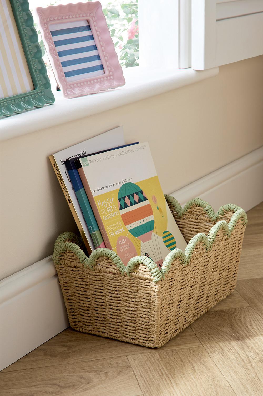 Scalloped edge woven basket with green edge, placed on the floor, holding multiple books. Pink and green photo frames on window sill.