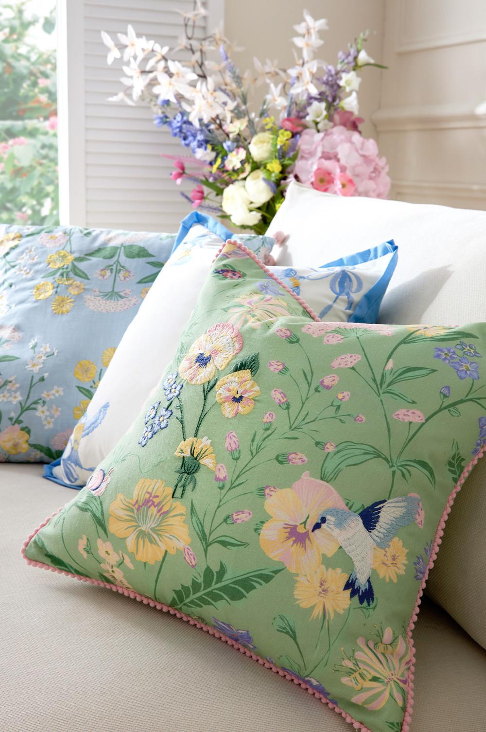 Floral cushion with mint green background featuring yellow flowers and blue hummingbird, trimmed with pink pom-pom edging.
