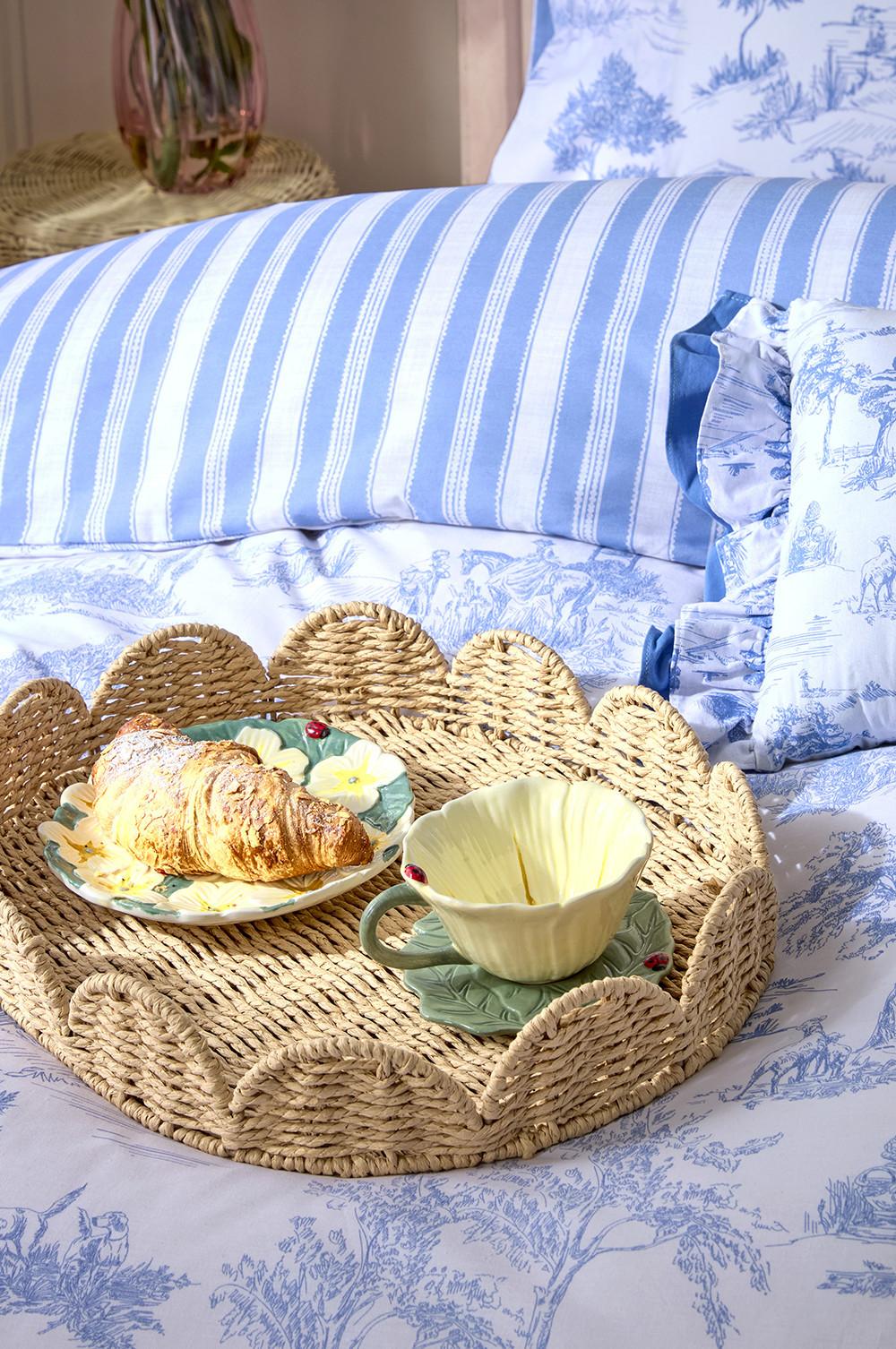 Oval, woven, scalloped tray, holding small flower printed plates and floral teacup and saucer. Tray is placed on white and blue patterned duvet.