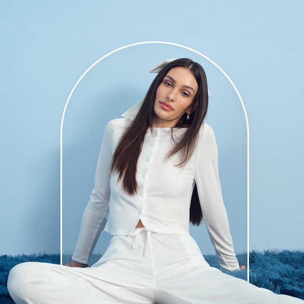 Model sitting on floor in white button-up long sleeve top, matching white pants, white hair bow and pearl earrings.
