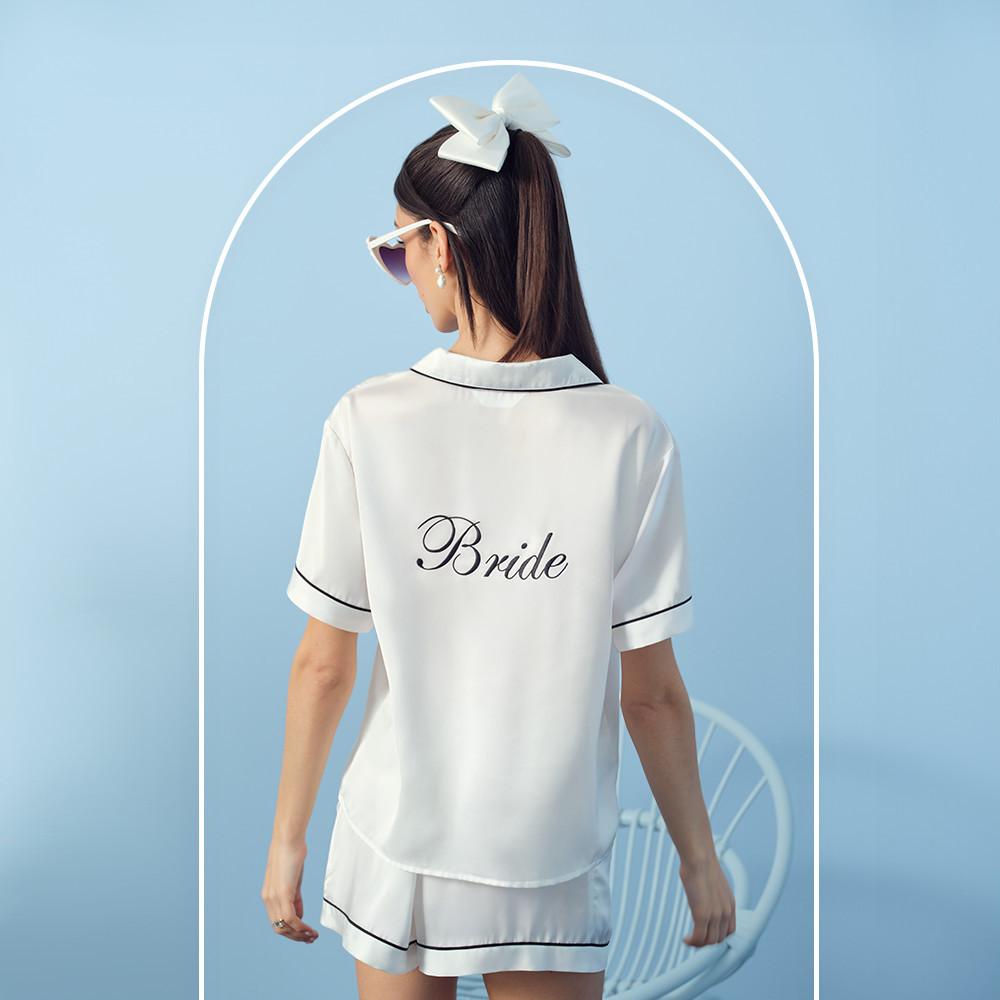Back of model in white shorts and shirt pajamas with black piping and Bride on the back. Hair worn with a white bow, with white sunglasses and pearl earrings.