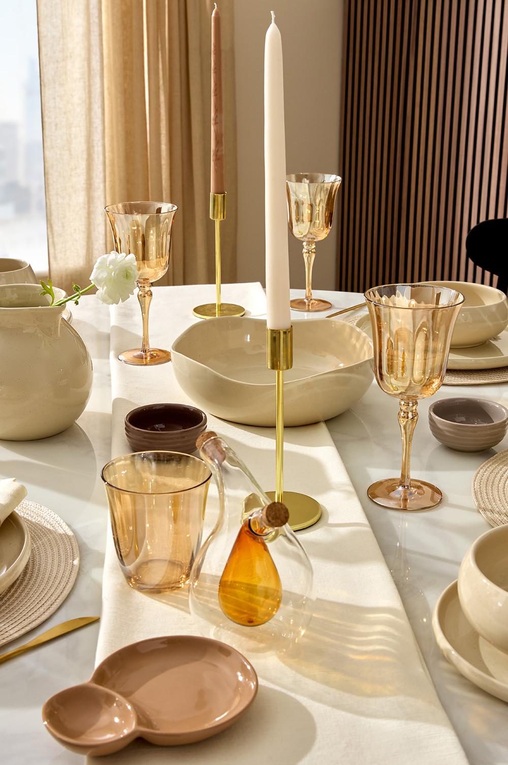 Elegante Tischdeko mit cremefarbenen Keramiktellern, gewebten Tischsets und bernsteingelben Gläsern auf weißer Tischdecke.