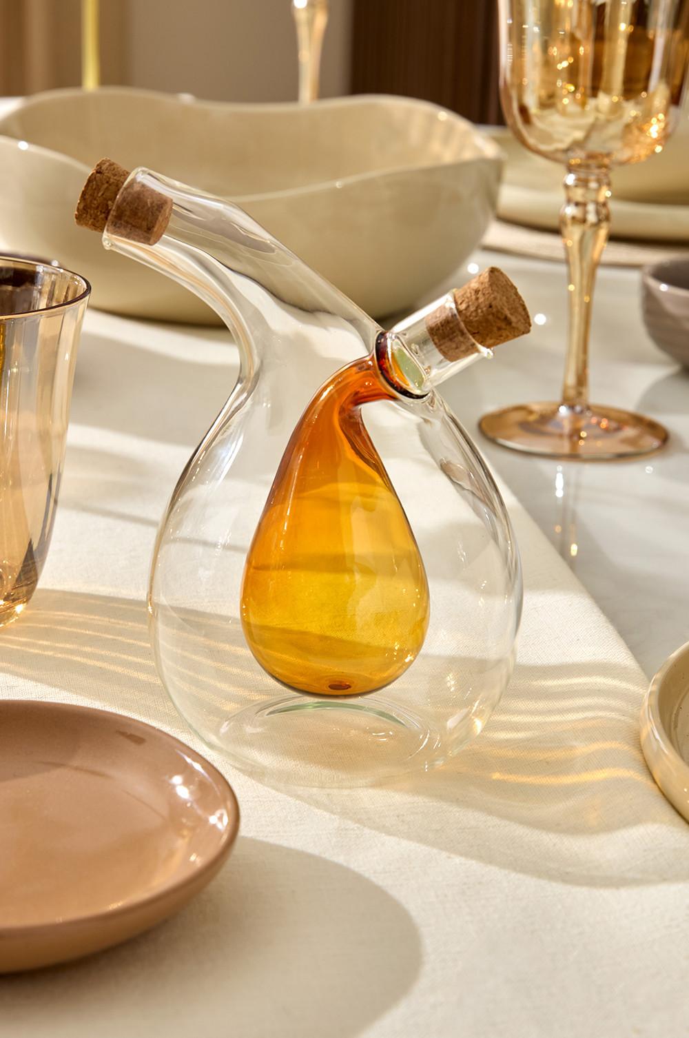 Glass oil and vinegar dispenser with amber-colored inner vessel and cork stoppers on white tablecloth.