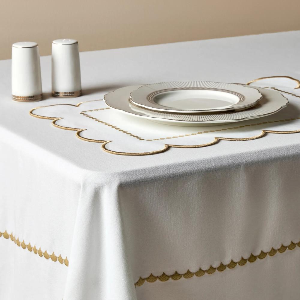 Table dressée élégamment avec des assiettes blanches, une salière et une poivrière sur une nappe blanche ornée d’une broderie festonnée dorée.
