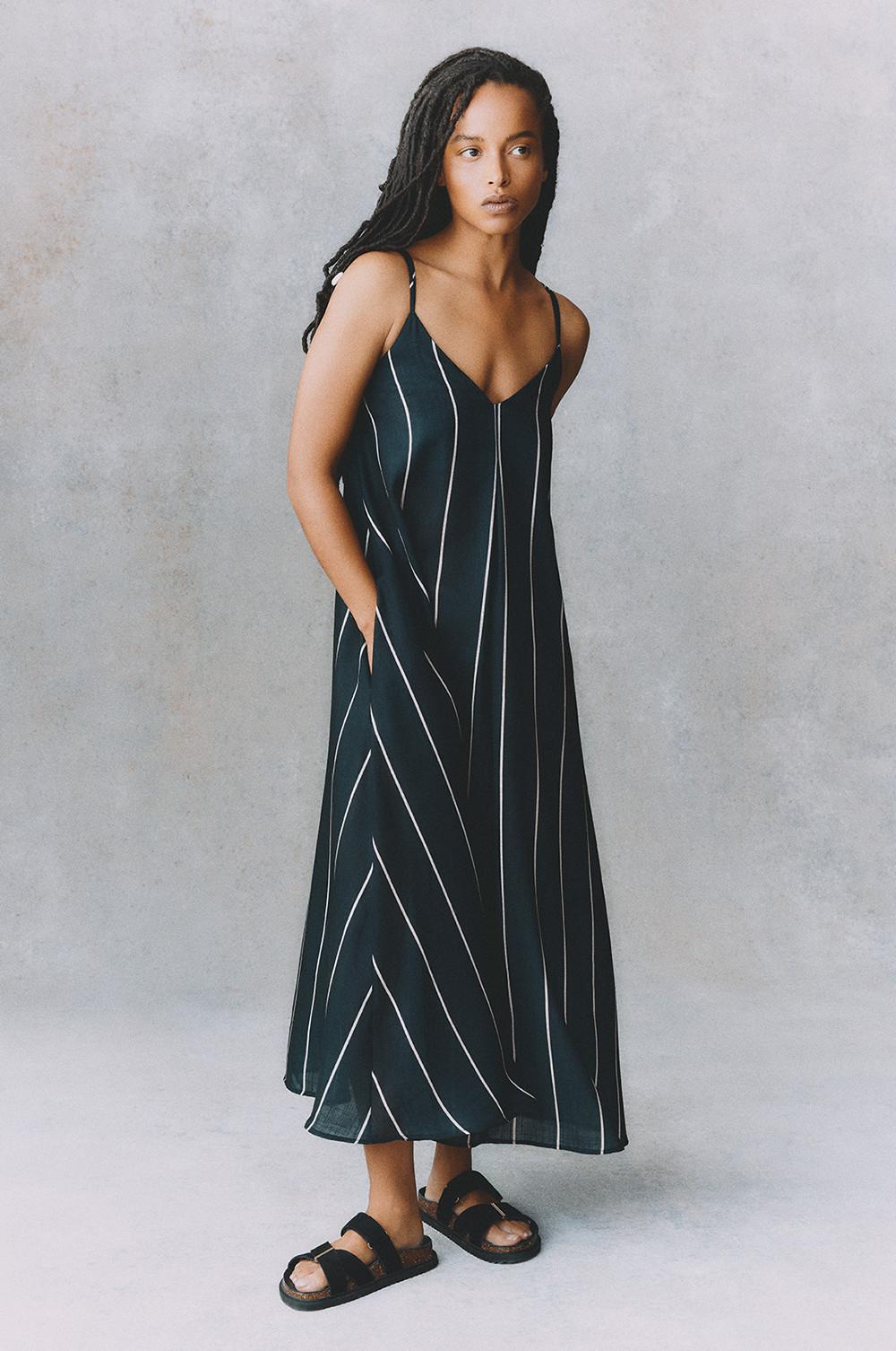 Model wearing navy maxi dress with white diagonal stripes, V-neckline and thin straps, paired with black sandals.