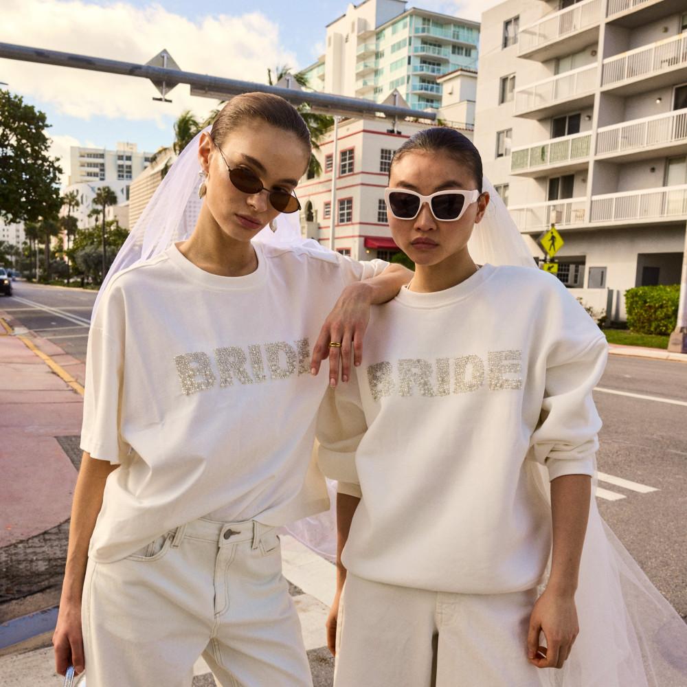 Two models wearing white BRIDE tops with veils and sunglasses, posing together on an urban street.