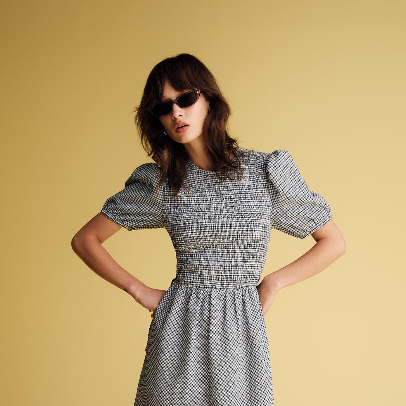 Model wearing black and white gingham dress with puffed sleeves and shirred bodice, styled with sunglasses.