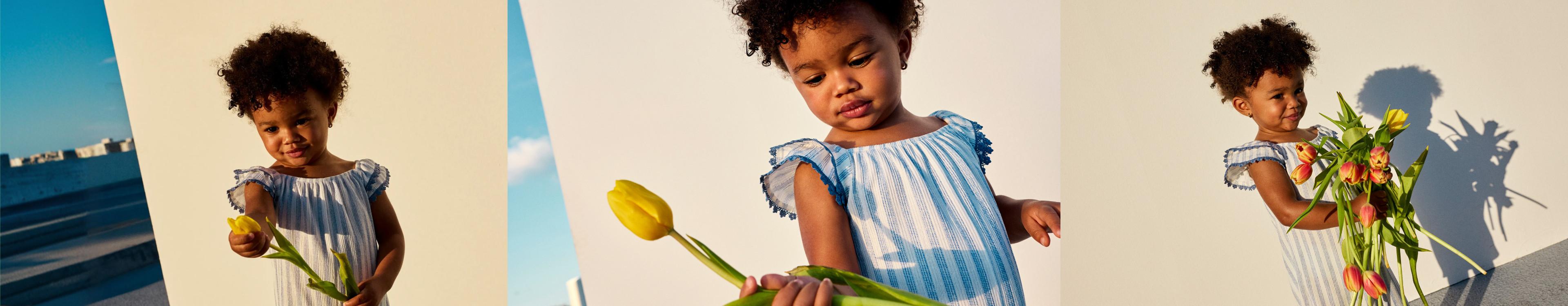Young child wearing a blue and white striped dress with pom-pom sleeves, holding yellow tulips.