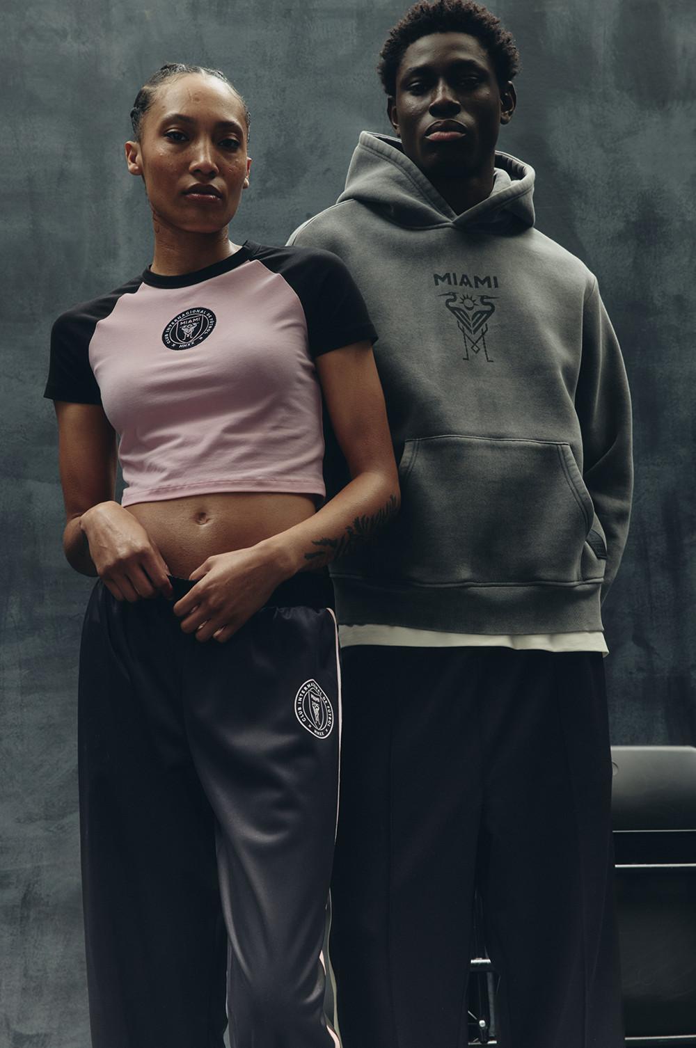 Zwei Models in „Inter Miami“ Sportbekleidung, darunter ein bauchfreies pinkes T-Shirt, eine graue Jogginghose, ein grauer Hoodie und eine dunkle Cargohose mit Teamlogos.