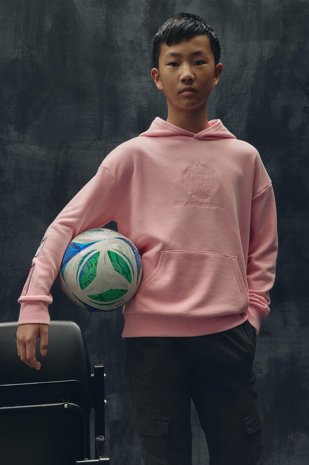 Ein Model im pinken Kapuzensweatshirt mit geprägtem Logo hält einen Fußball vor einem dunklen Hintergrund.