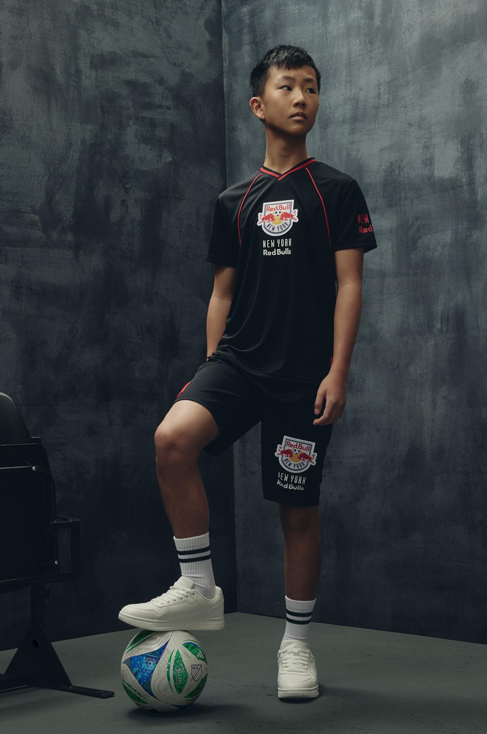 Schwarzer „New York Red Bulls“ Trainingsanzug mit rotem Besatz, dazu weiße Sneaker und ein Fußball, der zu Füßen des Models abgebildet ist.
