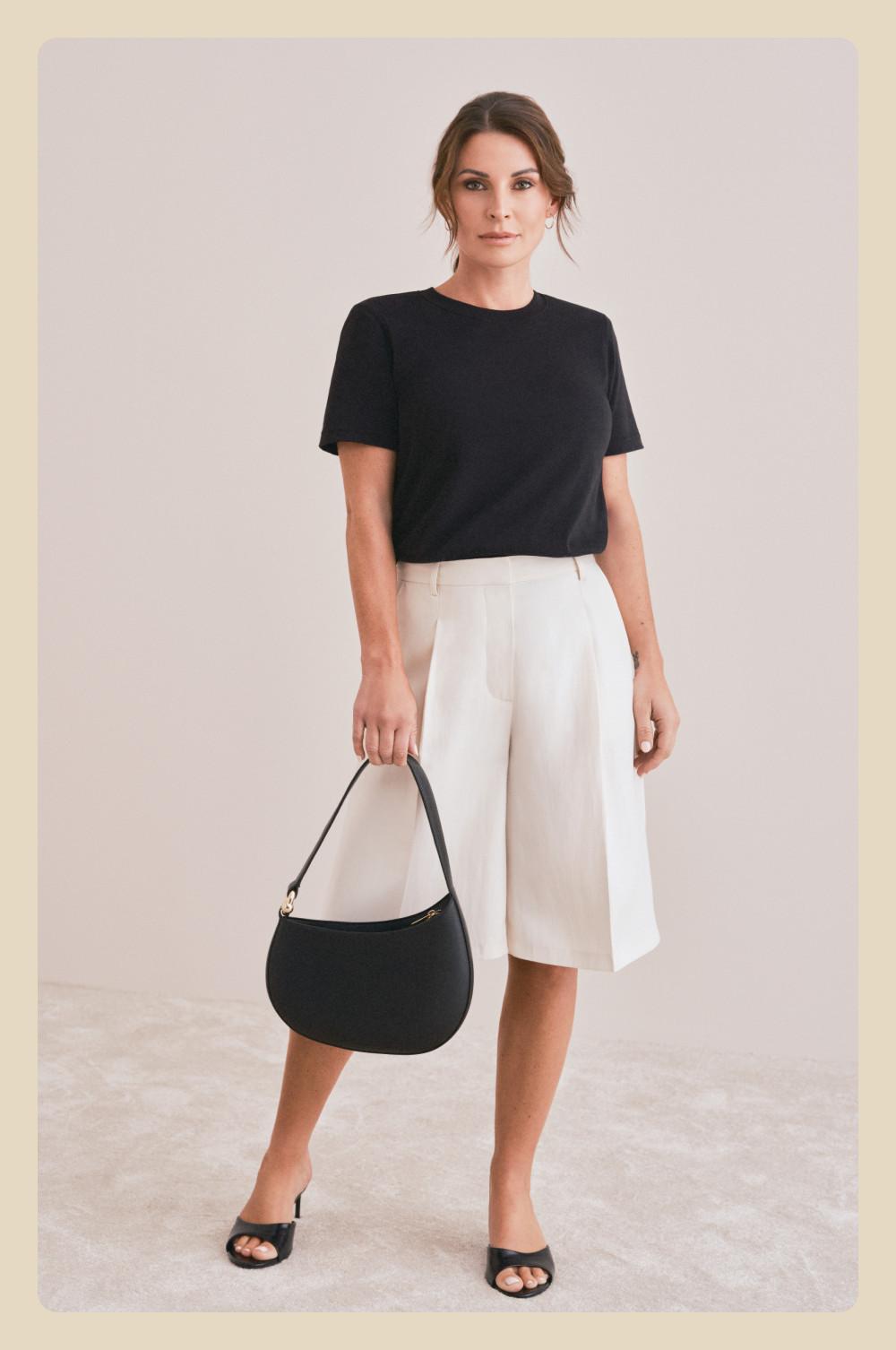Woman wearing black short-sleeved top and cream culottes, holding black shoulder bag, in black sandals.