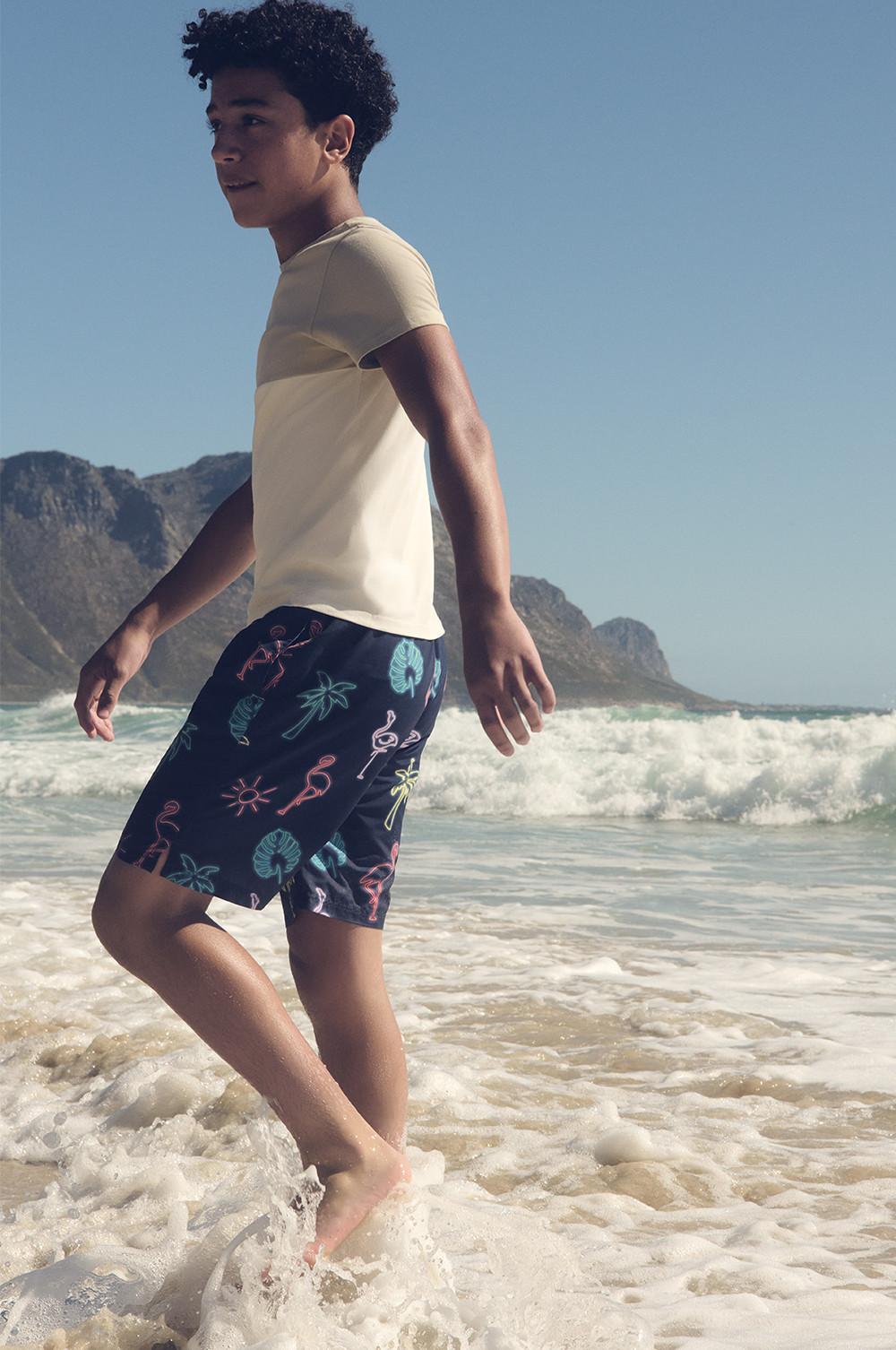 Kid wearing grey and cream T-shirt and navy swim shorts with tropical motifs walking on beach.