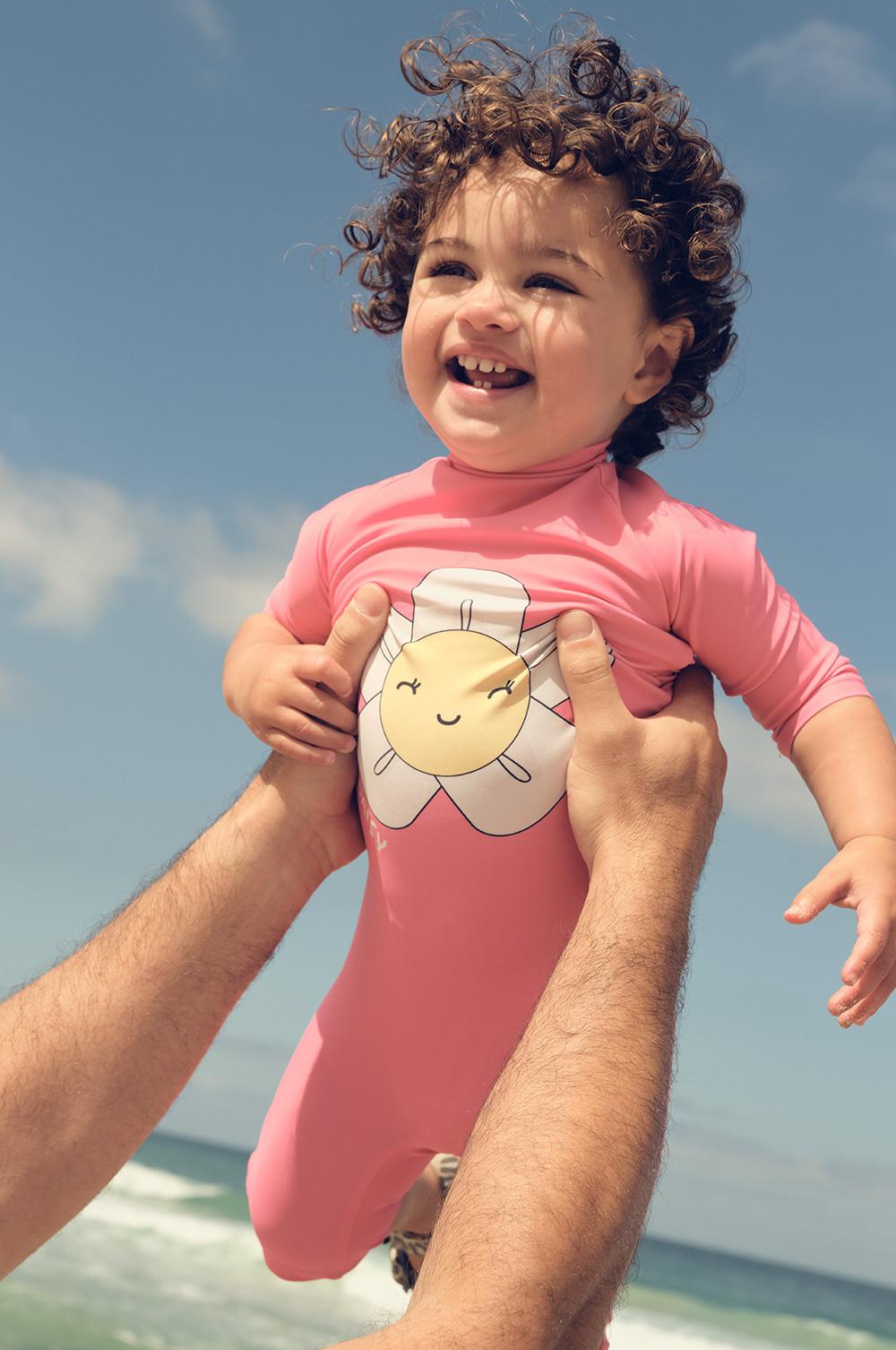 Child wearing pink sun protection swimsuit with smiling flower character design on chest, being held up at the beach against blue sky.