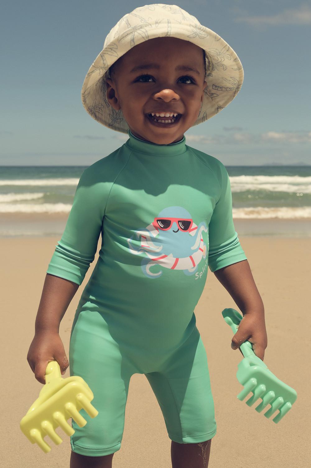 Child in mint green UV swimsuit with octopus graphic, wearing sun hat and holding beach sand toys at seaside.
