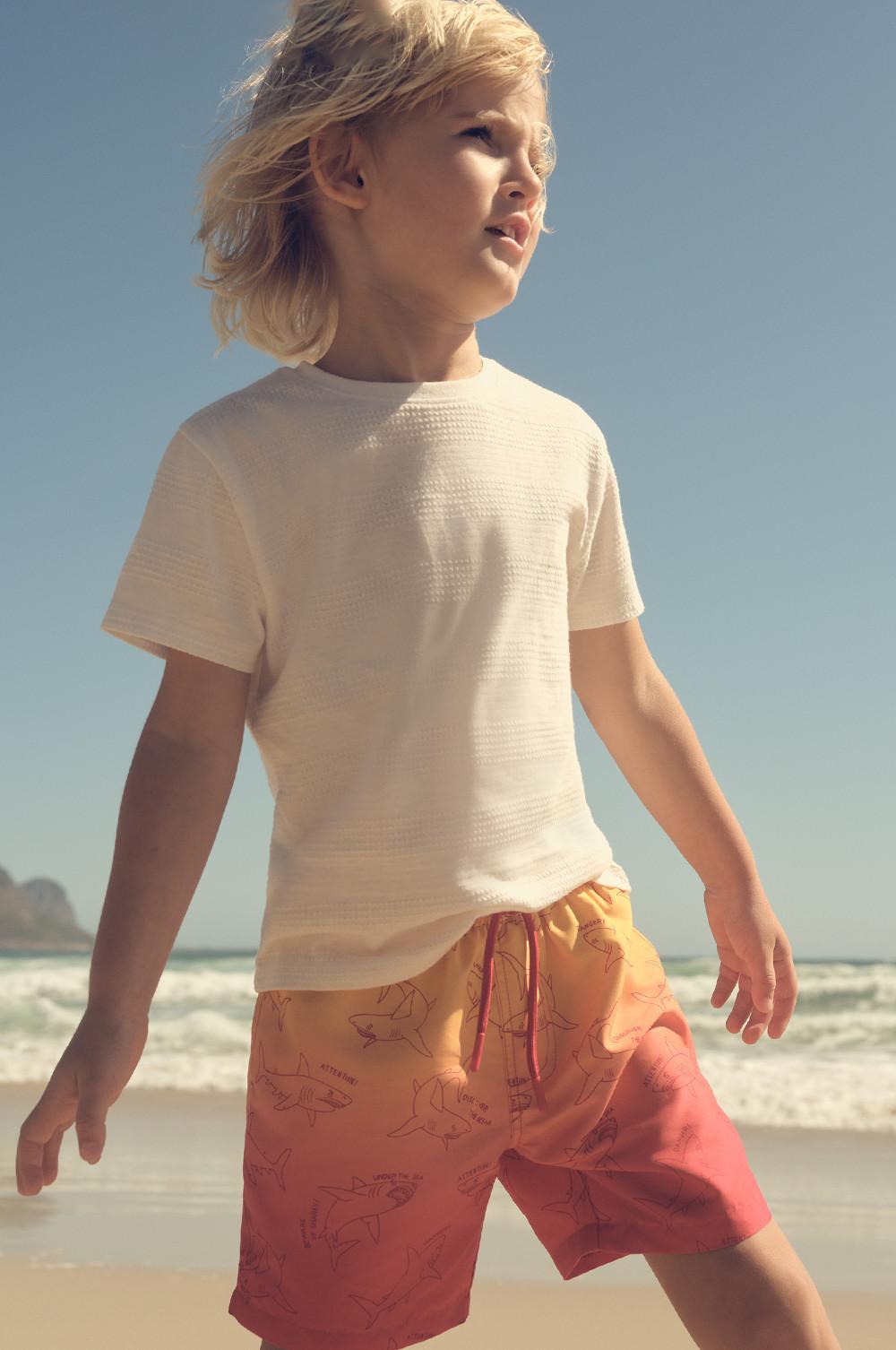 Child wearing white textured T-shirt and orange swim shorts with shark print pattern standing on beach.