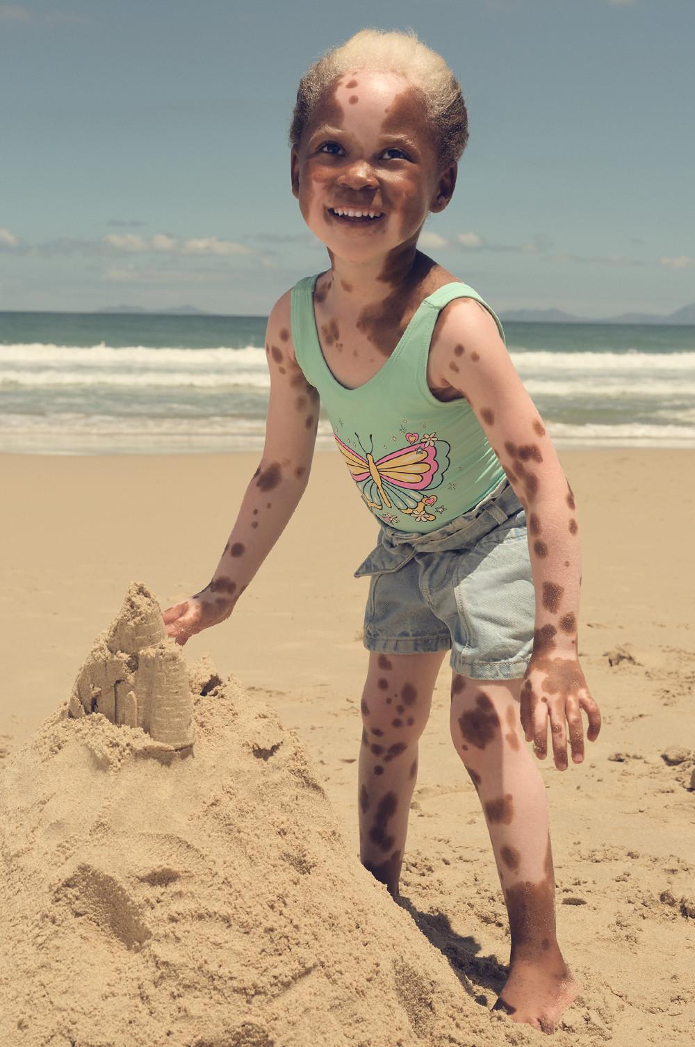 Child wearing mint green butterfly one-piece swimsuit and denim shorts building a sandcastle on sunny beach.