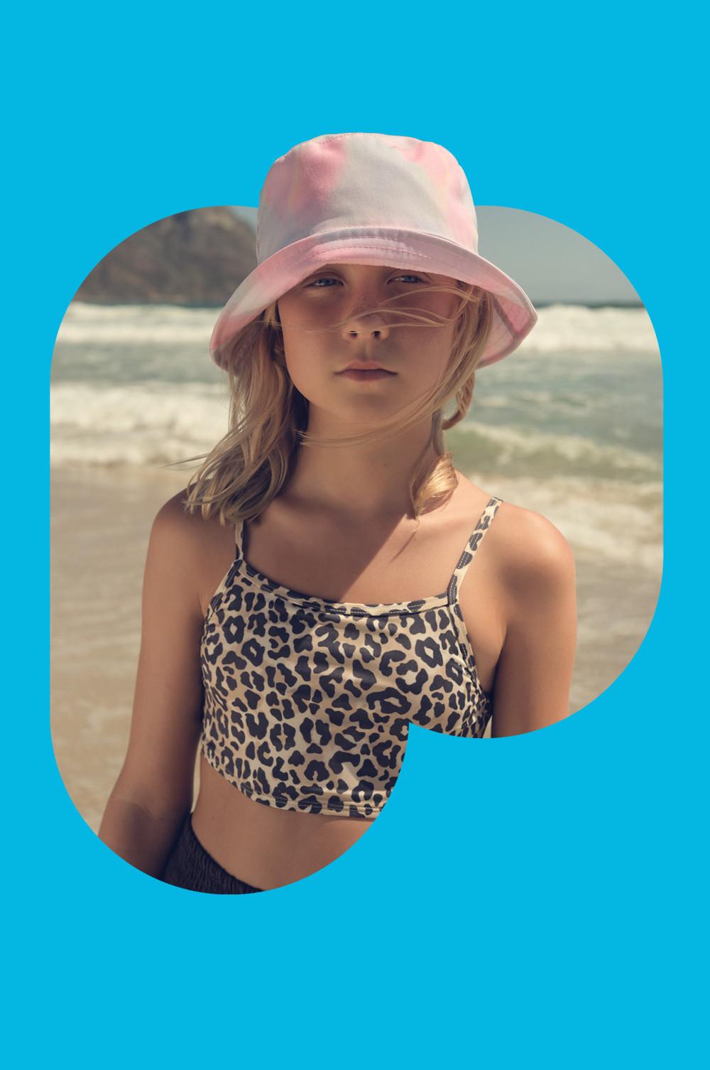 Leopard print crop top swimwear with thin straps and pink bucket hat worn at beach setting with waves visible.