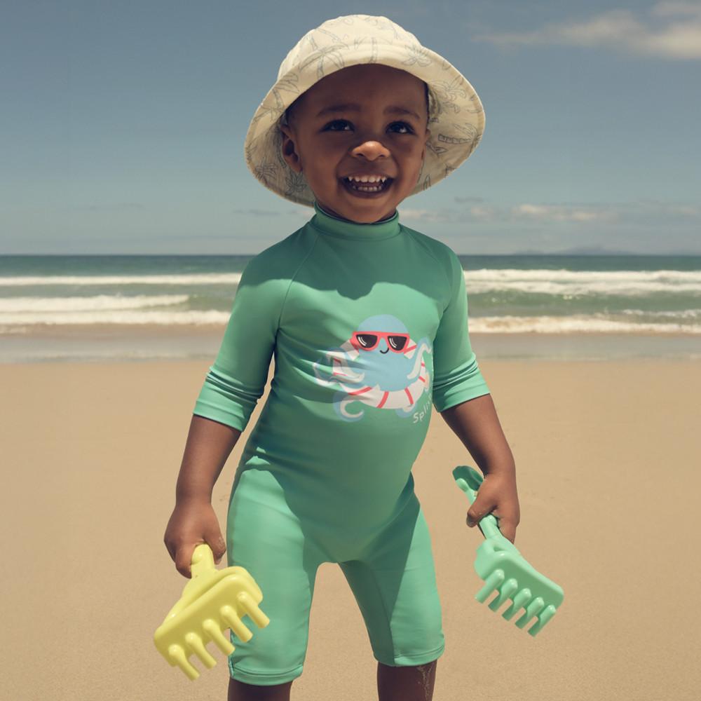 Child in mint green UV swimsuit with octopus graphic, wearing sun hat and holding sand toys at the beach.
