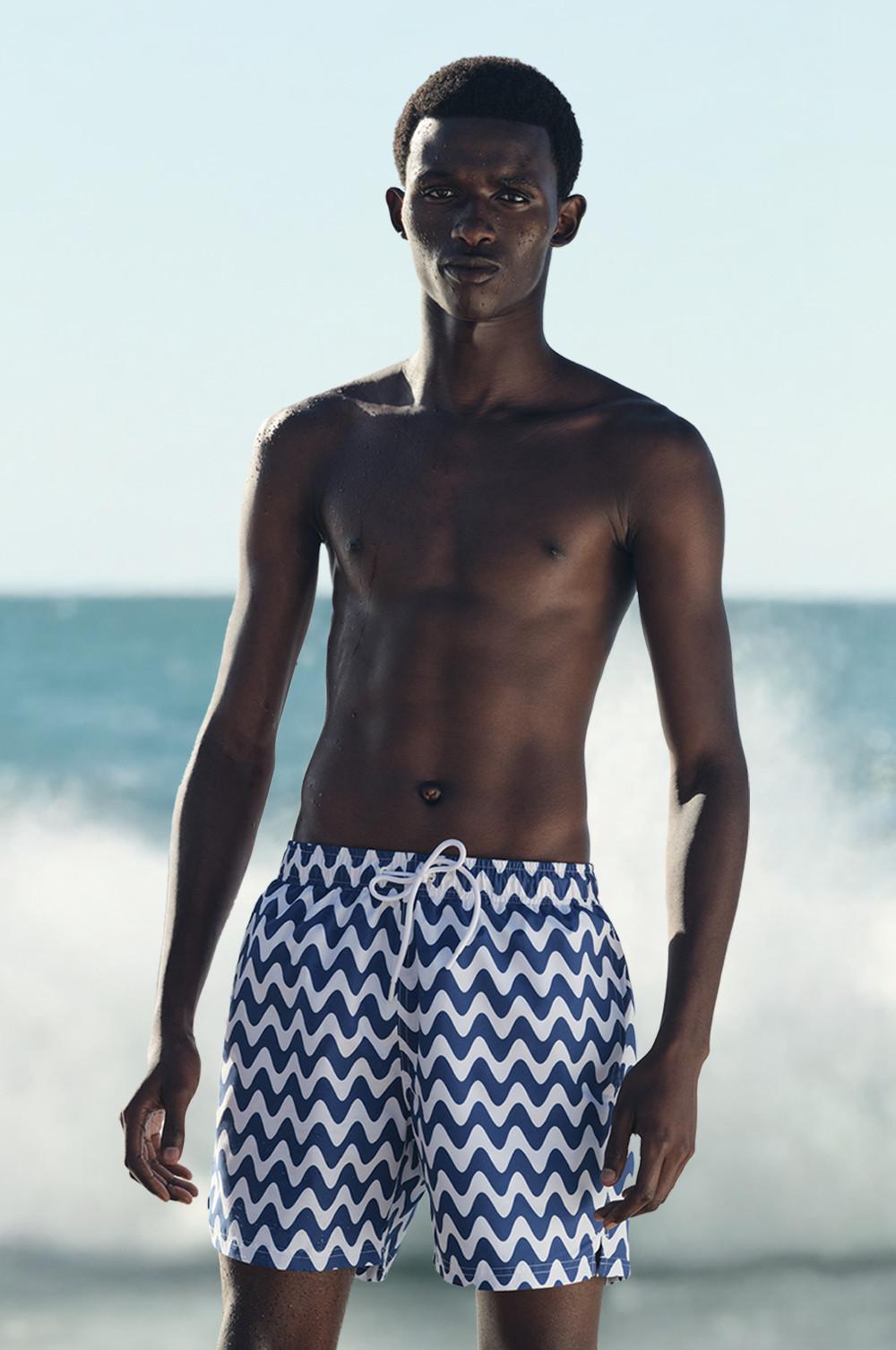 Costume da bagno con motivo a onde blu e bianche indossato da un modello davanti all'oceano con le onde che si infrangono.