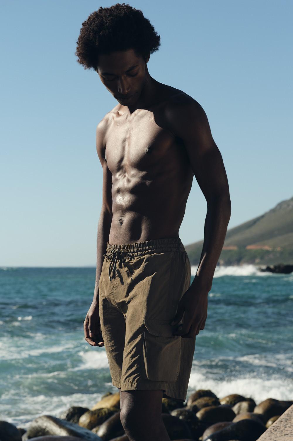 Persona a torso nudo che indossa pantaloncini da bagno cargo cachi, in piedi su una costa rocciosa con onde dell'oceano e scogliere sullo sfondo.