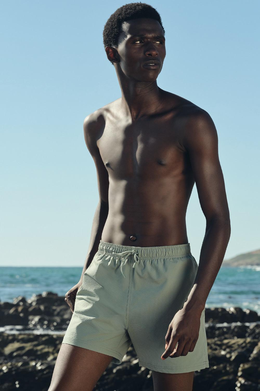 Modello che indossa pantaloncini da bagno verde menta, in piedi su una spiaggia rocciosa in riva all'oceano.